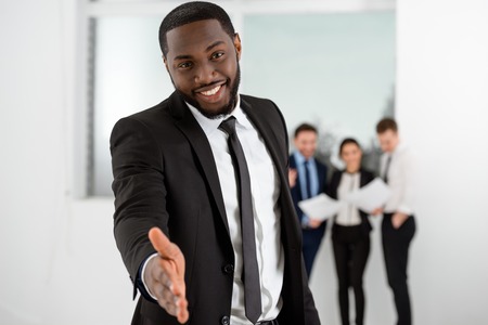 Young smiling African-american businessman looking at camera and reaching out his hand. Business people communicating in the backgroundの写真素材