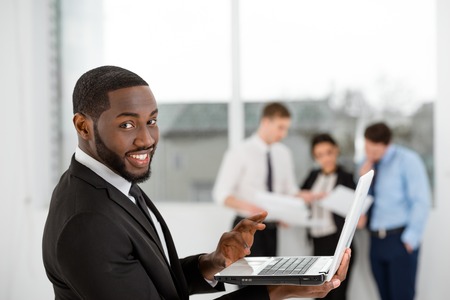 Young smiling African-american businessman looking at camera and using laptop. Business people communicating in the backgroundの写真素材