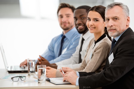 Cheerful multi-ethnic business team sitting in a row and lookingat camera. There are graphs and laptop on tableの写真素材