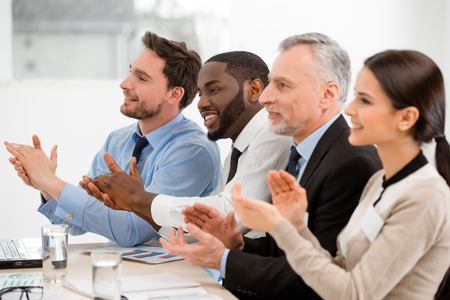Multi-ethnic business team sitting in a row, applauding and looking forward. There are graphs and laptop on tableの写真素材