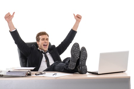 Photo of young cheerful businessman wearing suit. He sitting with his feet on desk with arms up. Isolated on white backgroundの写真素材