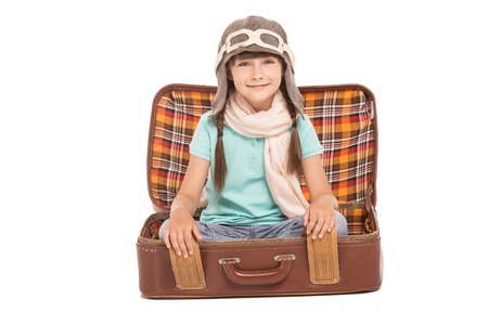 Cute little girl with two braids is isolated on white background. Girl sitting in vintage suitcase, looking at camera, smiling and wearing like a pilotの写真素材