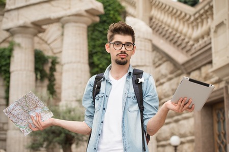 Portrait of handsome young man on vacation. Bewildered boy holding map and tablet computer. Boy looking at camera questioningly and trying to chooseの写真素材