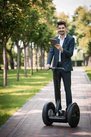 Portrait of handsome young businessman wearing suit. Man using segway and tablet computer. Man smiling and looking at camera. Green alley as backgroundの写真素材