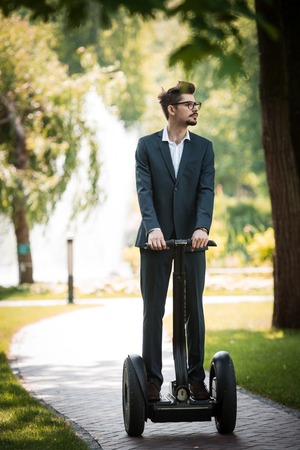 Portrait of handsome young businessman wearing suit. Man using segway and looking aside. Green alley...