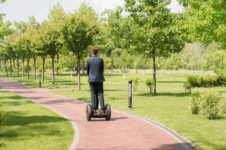 Back view of handsome young businessman wearing suit. Man using segway. Green alley as background
