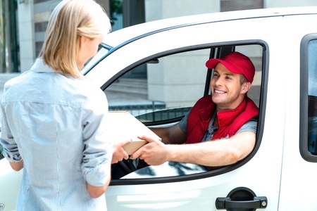 Colorful picture of courier delivers package for woman. Courier is sitting in the car and smiling. Woman accepts the parcel.の写真素材
