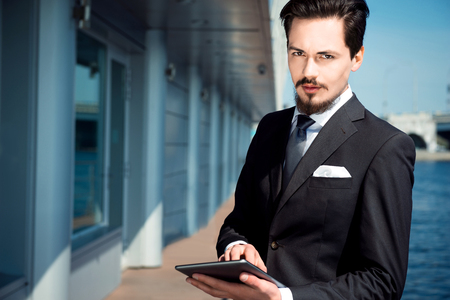 Photo of handsome young businessman standing near area river business centre. Man wearing suit, using tablet computer and looking at cameraの写真素材