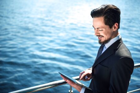 Photo of handsome young businessman standing near area river. Man wearing suit, smiling and using tablet computerの写真素材