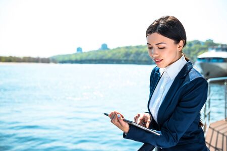 Photo of beautiful young business woman standing near area river. Woman wearing suit and using tablet computerの写真素材