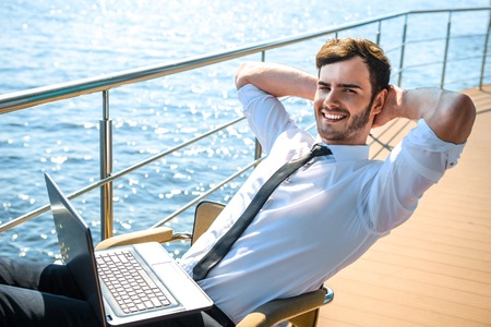 Photo of handsome young businessman sitting and relaxing near river. Man wearing suit, cheerfully smiling, looking at camera and using laptopの写真素材