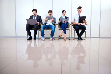 Photo of young creative business team of four with documents and computers. They sitting in a row and working. One man wearing informal clothesの写真素材