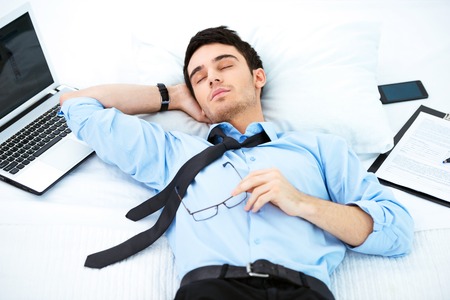 Young businessman napping in cozy hotel room on big white bed with laptop, documents and phone near himの写真素材