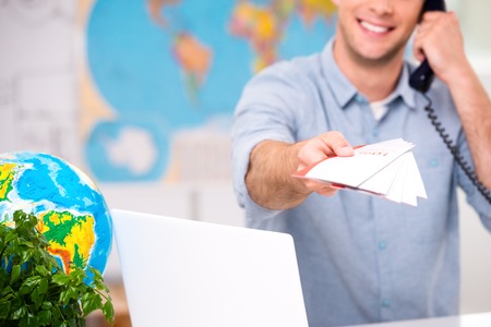Close up photo of male travel agent. Young man talking on phone, smiling, proposing tourist booklet and looking at camera. Travel agency office interior with big world map. Focus on bookletの写真素材