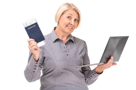 Portrait of senior blonde woman standing isolated on white background. Woman wearing grey blouse. Woman looking at camera, holding passport, air tickets, laptop and smilingの写真素材