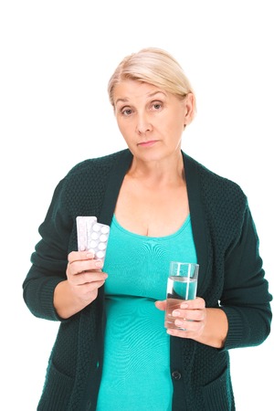 Portrait of senior blonde woman standing isolated on white background. Unhappy woman looking at camera, and holding pills and glass of waterの写真素材