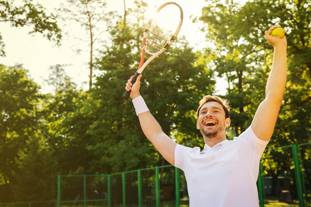 Picture of handsome young man on tennis court. Man playing tennis. Man cheerfully smiling and rejoicing after winning. Beautiful forest area as backgroundの写真素材