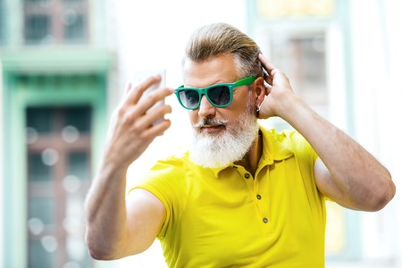 Portrait of stylish handsome adult man with beard standing outdoors. Man wearing glasses and yellow T-shirt, and making selfie photo with mobile phoneの写真素材
