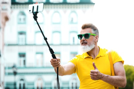 Portrait of stylish handsome adult man with beard standing outdoors. Man wearing glasses and yellow T-shirt, showing thumb up and making photo with selfie stickの写真素材