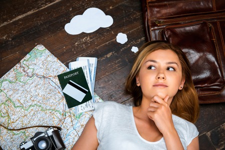 Top view photo of beautiful blonde girl lying on wooden floor. Young woman with thoughts on cloud. Passport, tickets, vintage camera, credit card and map are on floorの写真素材
