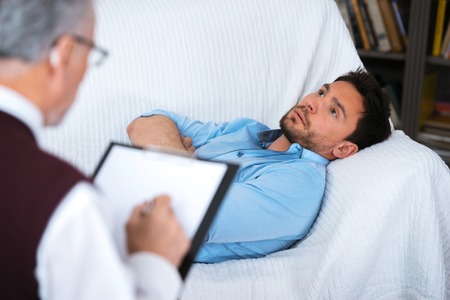 Sad young man talking with psychologist and lying on coach. Psychologist taking notes. There are many books in psychologist officeの写真素材