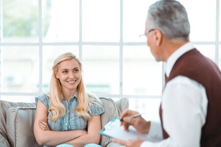 Smiling happy young woman talking with psychologist. Psychologist taking notes. There is big window in psychologist officeの写真素材