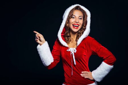 Christmas portrait of beautiful mixed-race santa girl. Young sexy brunette woman wearing santa claus clothes. Snow Maiden pointing aside on black backgroundの写真素材