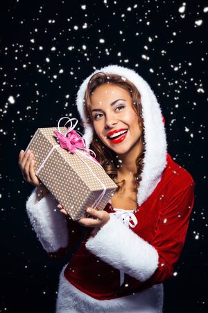 Christmas portrait of beautiful mixed-race santa girl. Young sexy brunette woman wearing santa claus clothes. Snow Maiden holding present on black background with snowの写真素材