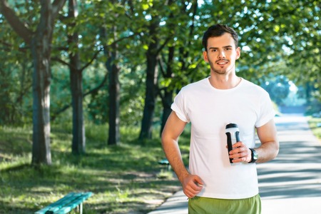 Photo of handsome nice guy outdoors at morning. Young man looking at camera and holding sport drink...
