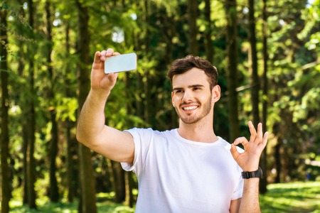 Photo of handsome nice guy outdoors at morning. Young man smiling, showing ok sign and making selfie photo on mobile phone while running in parkの写真素材