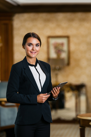 Young business woman wearing suit, standing in nice hotel room, using tablet computer and looking at cameraの写真素材