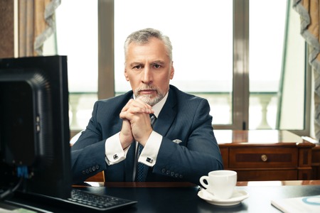 Photo of businessman in expensive and classic office. Aged businessman wearing suit, sitting near computer, having coffee and looking at cameraの写真素材