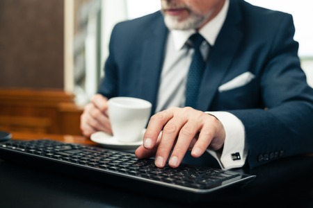 Aged businessman wearing suit, using computer and having coffeeの写真素材