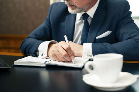 Close up photo of businessman in expensive and classic office wearing suit, making notes and having coffeeの写真素材