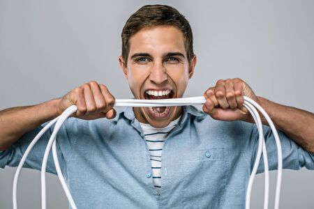 Portrait of handsome stylish young man standing on grey background. Angry man with wires on hands trying to bite itの写真素材