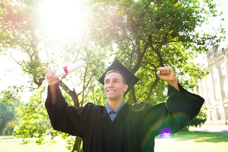Young male student dressed in black graduation gown. Campus as a background. Boy cheerfully smiling with arms up, holding diploma and looking aside. Light flare on photoの写真素材