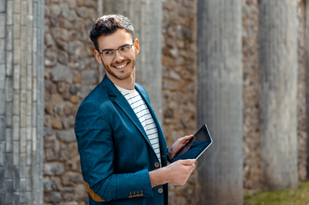 Portrait of stylish handsome young man with bristle standing outdoors. Man wearing jacket and shirt. Smiling man with glasses holding tablet computerの写真素材
