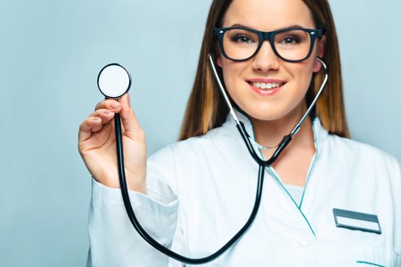 Portrait of female doctor with glasses, stethoscope and lab coat. Young smiling doctor holding stethoscope and looking at camera. Woman standing on grey backgroundの写真素材