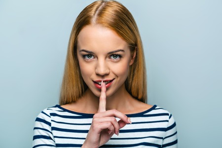 Portrait of beautiful caucasian blonde woman standing on grey background. Young woman looking at camera and making silence signの写真素材