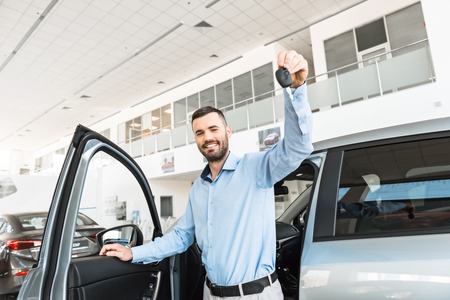 Photo of happy young man coming out of new car and showing keys to it. Concept for car rental