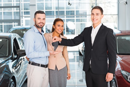 Photo of young male consultant presenting new car to buyers and giving them keys in auto show....