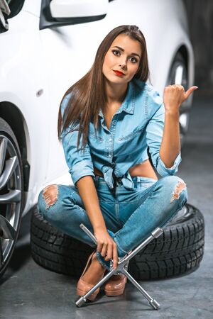 Photo of young female car repair worker. Glamour sexy brunette wearing jeans. Girl with wrench looking at camera, lsitting on tire near car and making hitchhiking gestureの写真素材