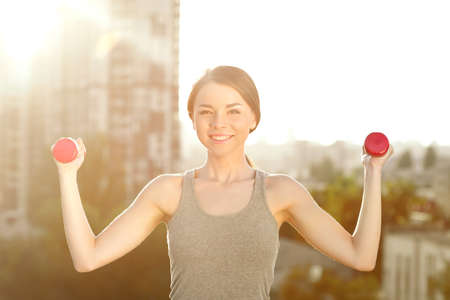Young woman practicing yoga on the mat on the roof, she holding dumbbells and looking at camera