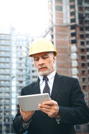 Business portrait of contractor and developer. Aged businessman with helmet standing near his new construction and using tablet computerの写真素材
