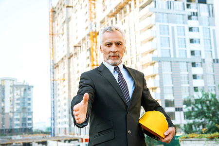 Business portrait of contractor and developer. Aged businessman with helmet standing near his new construction. Man proposing hand to shake and looking at cameraの写真素材