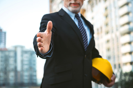 Business portrait of contractor and developer. Aged businessman with helmet standing near his new construction. Man proposing hand to shake at cameraの写真素材