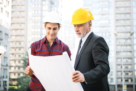 Portrait of engineer and builder. Men with helmets standing near new unfinished construction and discussing graphics and blueprintsの写真素材