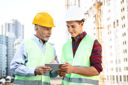 Portrait of contractor and builder in uniforms. Men with helmets standing near new unfinished construction and using tablet computerの写真素材