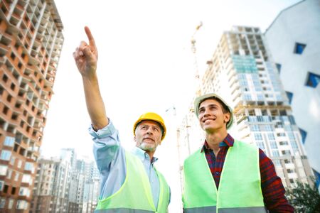 Portrait of contractor and builder in uniforms. Men with helmets standing near new unfinished construction. Aged man pointing somewhereの写真素材