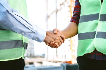 Portrait of contractor and builder in uniforms. Men standing near new unfinished construction and shaking handsの写真素材